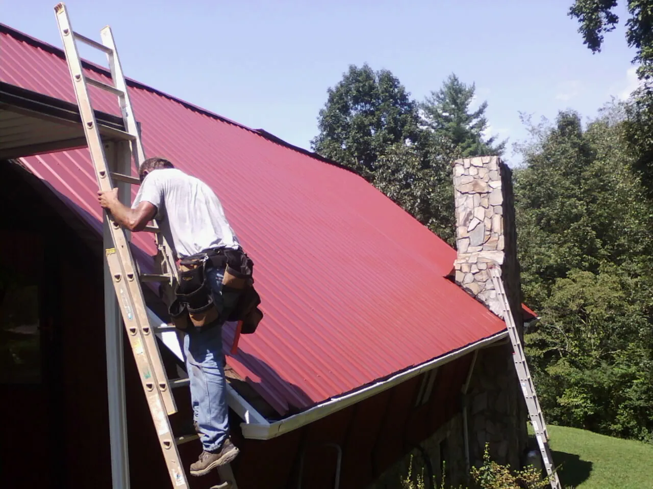 Roofer on a ladder beside a red metal roof and stone chimney — Soffit & Fascia Repair by Roofing Brothers in Waterville Valley